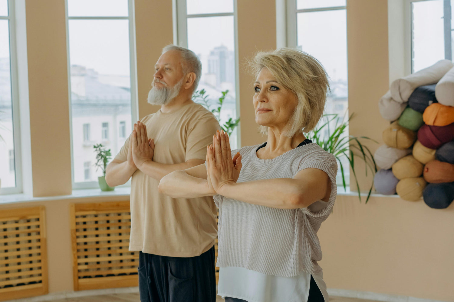 Two people practicing yoga in a room with large windows and a colorful wall hanging.